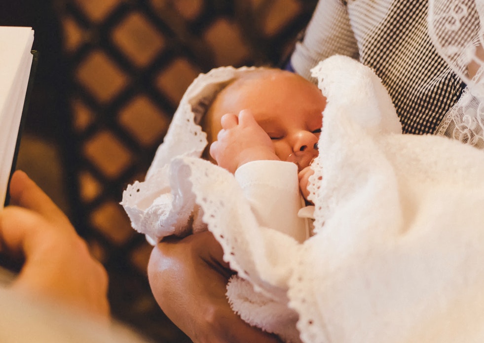 Das Foto wurde über die Schulter des Priesters gemacht, deshalb sieht man einen Teil der Bibel und davor ein Baby in den Armen seiner Mutter. Besondere Momente bedürfen besonderen Geschenken. Mit Golden Gates Gold schenken.