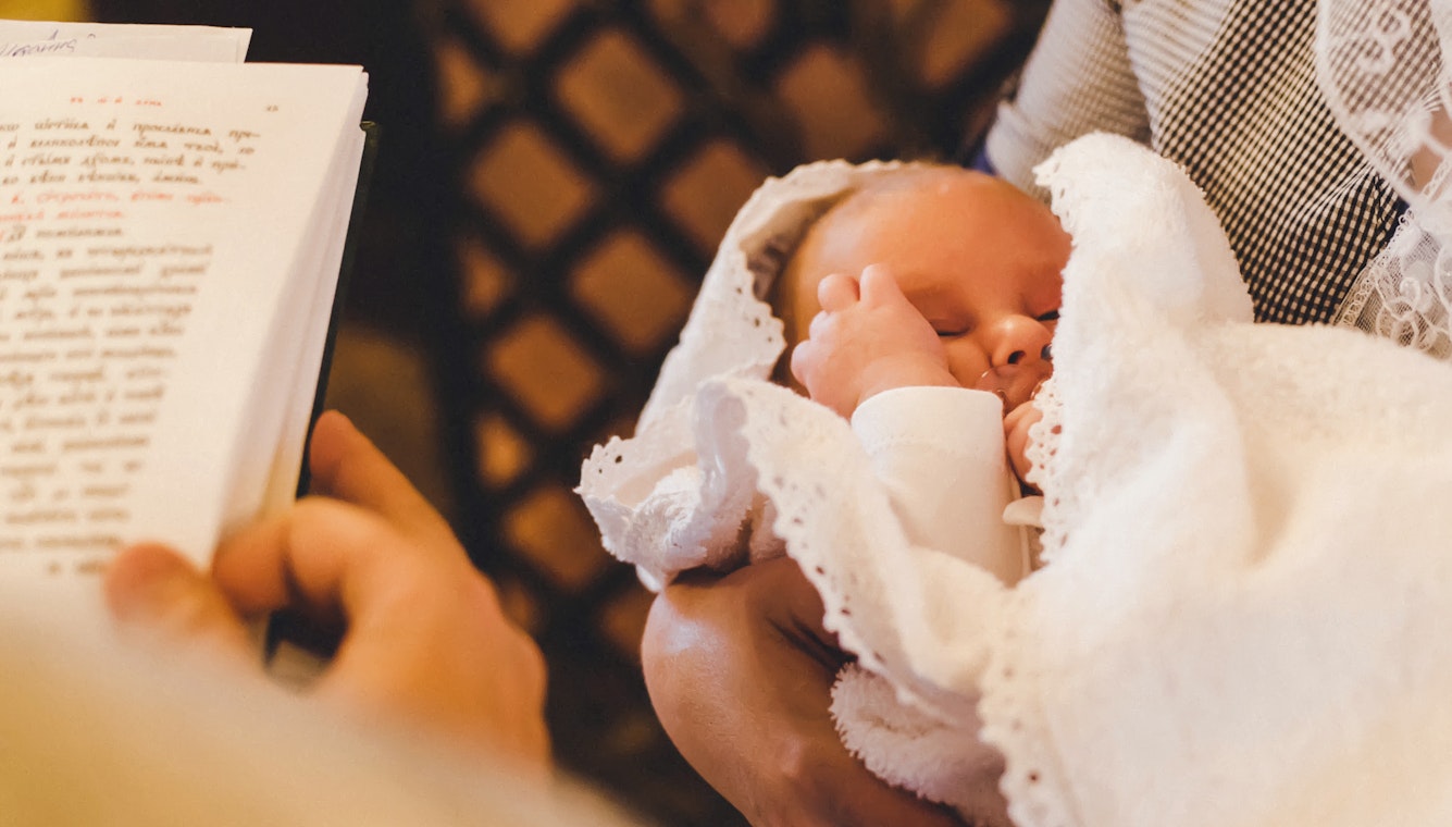 Das Foto wurde über die Schulter des Priesters gemacht, deshalb sieht man einen Teil der Bibel und davor ein Baby in den Armen seiner Mutter. Besondere Momente bedürfen besonderen Geschenken. Mit Golden Gates Gold schenken.