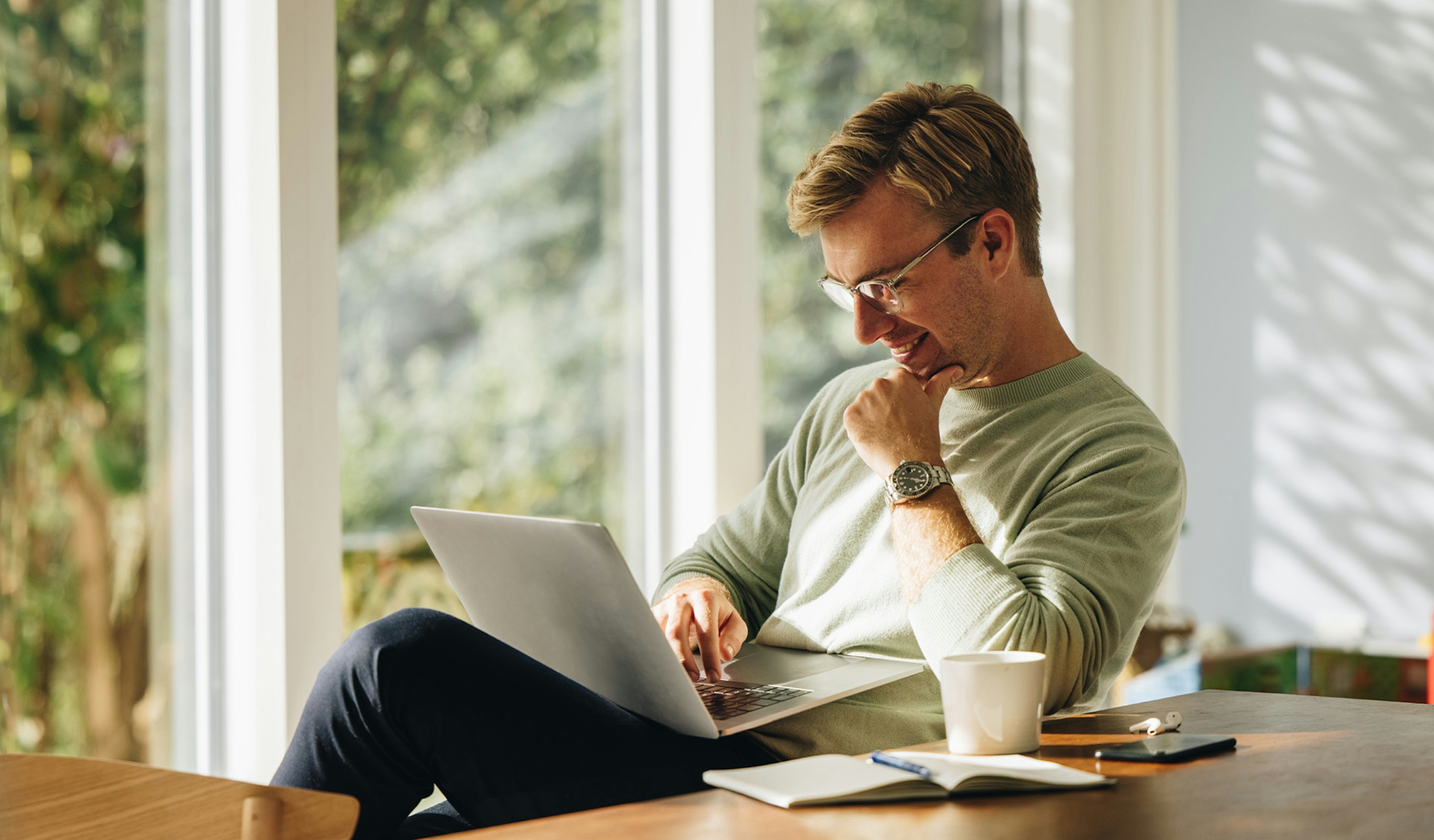 In einen hellen Raum, sitzt ein Mann an einem Tisch und blickt lächelnd auf den Laptop auf seinem Schoß. Entdecken auch Sie die Sparpläne von Golden Gates und legen Ihr Geld in Edelmetalle an.